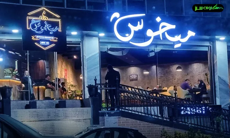 این تصویر نمای بیرونی رستوران میخوش را در شب نمایش می‌دهد. تابلوی نئونی آبی با نام رستوران، فضای داخلی با نورپردازی گرم و میزهای پر از مشتریان را نشان می‌دهد.